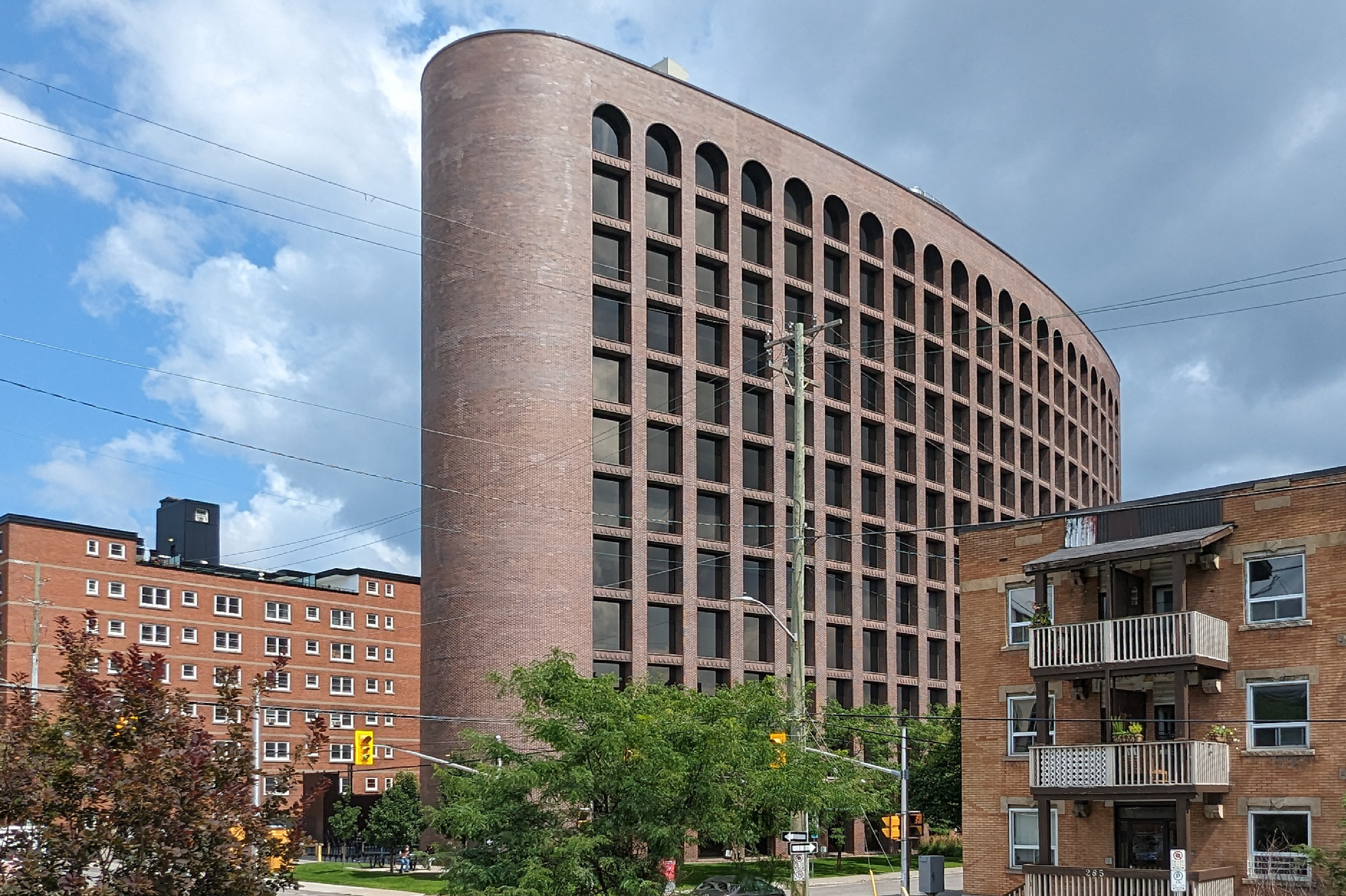 Tall and wide brown brick building in the shape of an extruded ellipse with curved walls and an orderly grid of square punch windows. The top row of windows is arched with a semicircle. Around it are 4 to 5 storey high brick buildings, rectangular in shape.